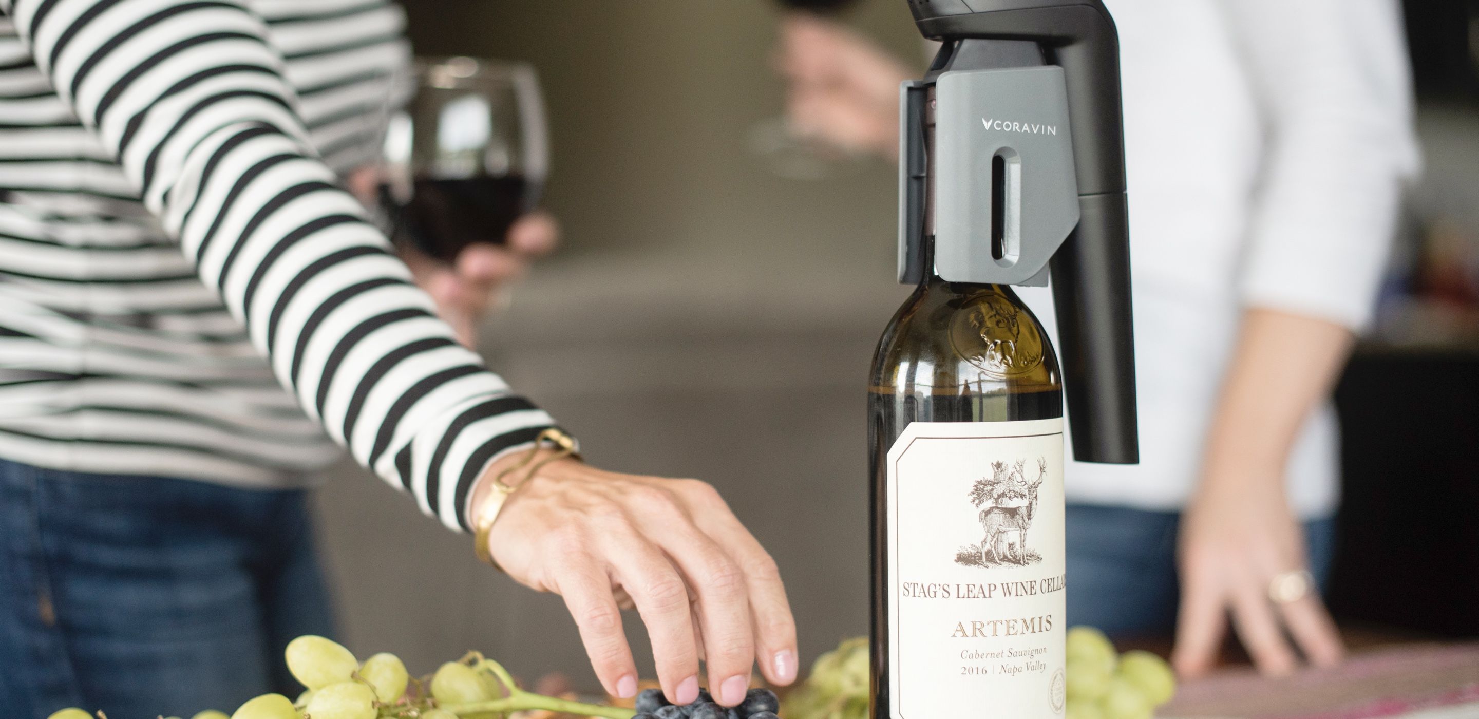 Coravin wine preservation system on top of a wine bottle. A person is grabbing grapes from beside the bottle.